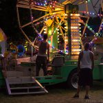 two men dismantling a childrens ride at a county fair, in the evening with lights